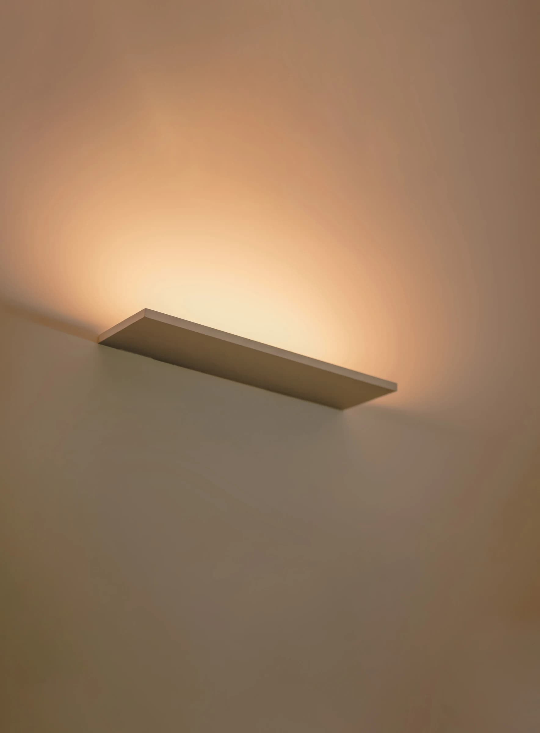 Minimal wall-wash shelf light casting warm upward glow on plaster wall
